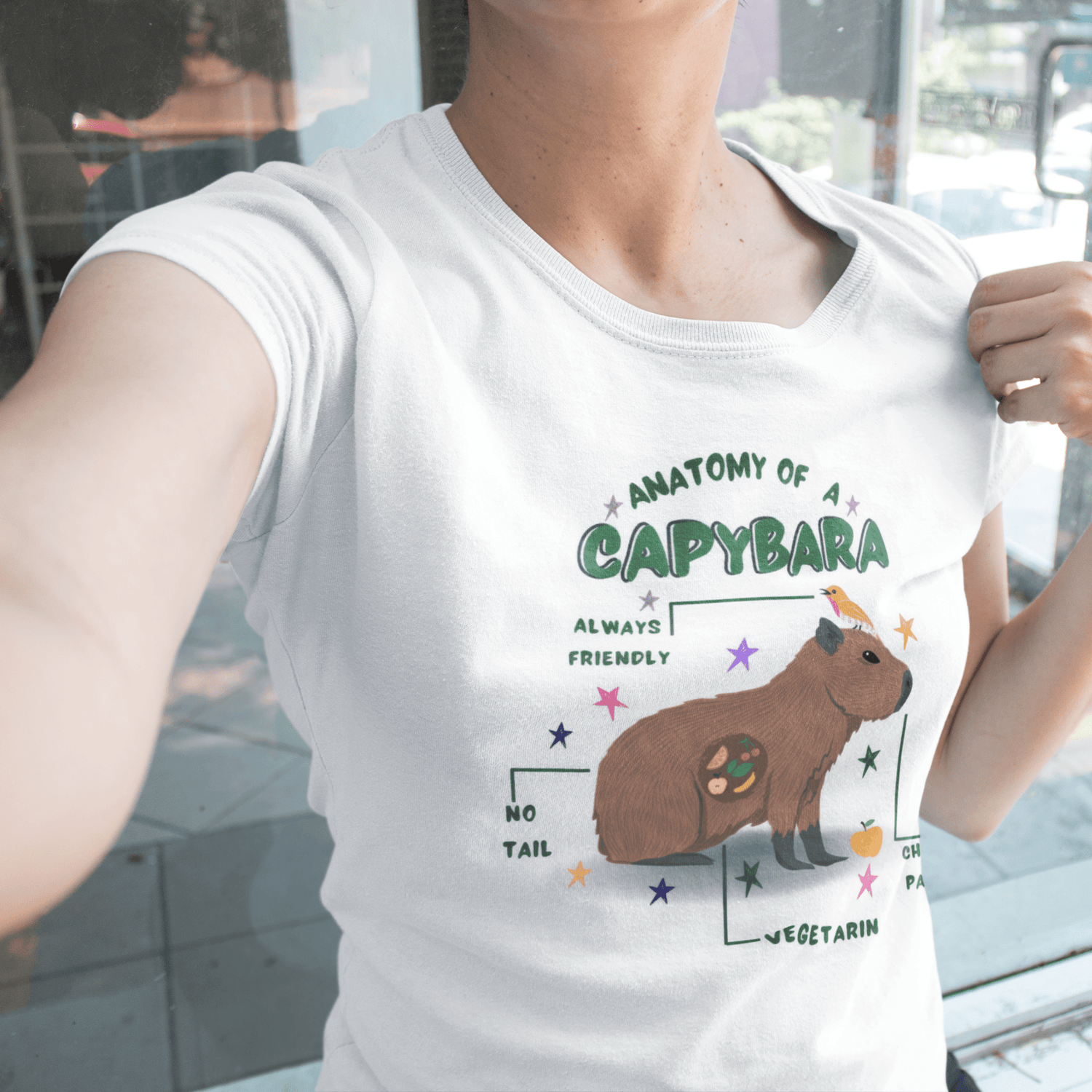 A person wearing a white graphic t-shirt with a colorful 'Anatomy of a Capybara' print, photographed in a casual selfie-style setting.